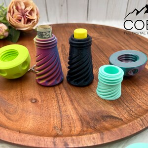 May include: A collection of 3D printed screw-top containers in various colors including lime green, a purple and yellow gradient, solid black, and mint green. One container holds a rolled twenty-dollar bill. The COBO The Digital File Shop logo is visible.