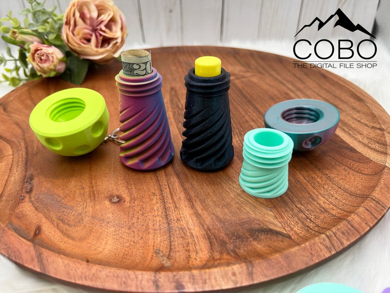 May include: A collection of 3D printed screw-top containers in various colors including lime green, a purple and yellow gradient, solid black, and mint green. One container holds a rolled twenty-dollar bill. The COBO The Digital File Shop logo is visible.