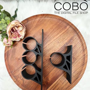 May include: Two black metal shelf brackets with a geometric design. The bracket on the left has three circular cutouts. The bracket on the right has one circular cutout. The brackets are on a wooden surface.