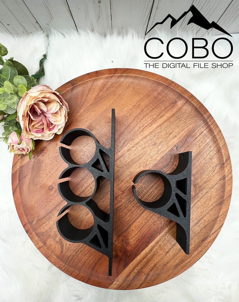 May include: Two black metal shelf brackets with a geometric design. The bracket on the left has three circular cutouts. The bracket on the right has one circular cutout. The brackets are on a wooden surface.