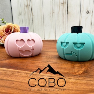 May include: Two 3D-printed pumpkin decorations. One is light pink with heart-shaped eyes and a purple stem. The other is teal with an angry face and a black stem. The pumpkins are on a wooden surface.