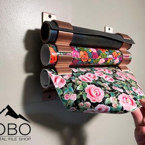 May include: A wall-mounted copper-colored metal holder with three rolls of wrapping paper. The top roll is black, the middle roll has a floral pattern with pink, orange, and green flowers, and the bottom roll has a floral pattern with pink flowers on a black background.