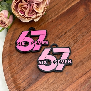 May include: Two pink and black charms with the number 67 and the words "SIX" and "SEVEN" printed on them. The charms are on a wooden surface, with a rose in the background.