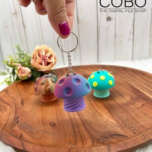 May include: Three mushroom-shaped keychains with metal rings and chains. The central keychain features a purple and blue gradient cap with circular indentations and a spiral stem. Two additional keychains are visible in the background.