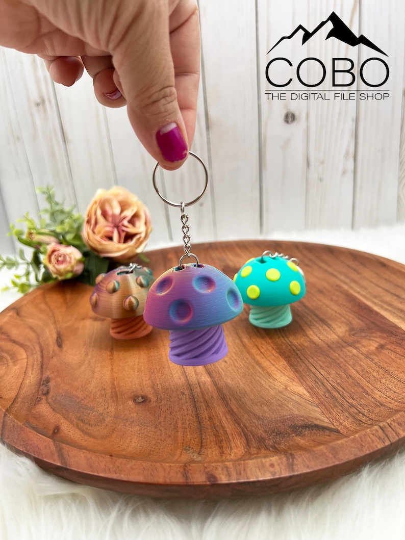 May include: Three mushroom-shaped keychains with metal rings and chains. The central keychain features a purple and blue gradient cap with circular indentations and a spiral stem. Two additional keychains are visible in the background.