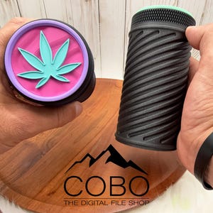 May include: Two black cylindrical containers, one with a teal cannabis leaf design on a pink and purple lid, and the other with a spiral pattern. The containers are held over a wooden tray with the text "COBO THE DIGITAL FILE SHOP".