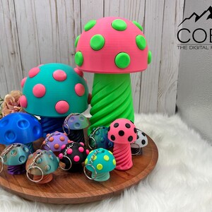 May include: A collection of 3D printed mushroom figures and keychains in various sizes and colors, including a large pink mushroom with green spots, a teal mushroom with pink spots, and smaller mushroom keychains in blue, purple, and black.