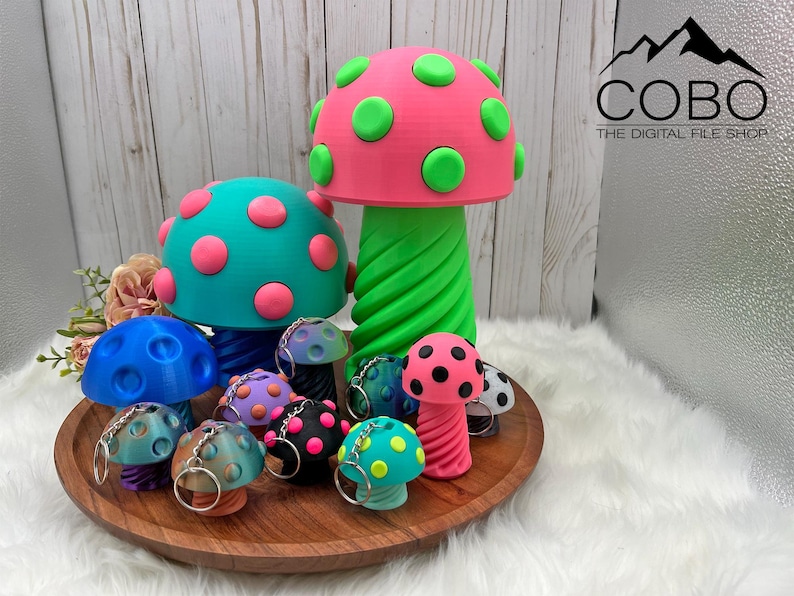 May include: A collection of 3D printed mushroom figures and keychains in various sizes and colors, including a large pink mushroom with green spots, a teal mushroom with pink spots, and smaller mushroom keychains in blue, purple, and black.
