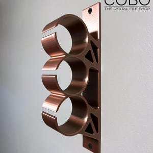 May include: A copper-colored metal wall mount with three circular openings for holding items. The mount is attached to a white wall with screws. The text "COBO THE DIGITAL FILE SHOP" is visible in the upper right corner of the image.