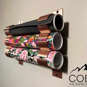 May include: A copper wall-mounted organizer with three rolls of patterned wrapping paper. The organizer has a corrugated design and is mounted to a wall with screws. The rolls of wrapping paper are in various floral patterns.