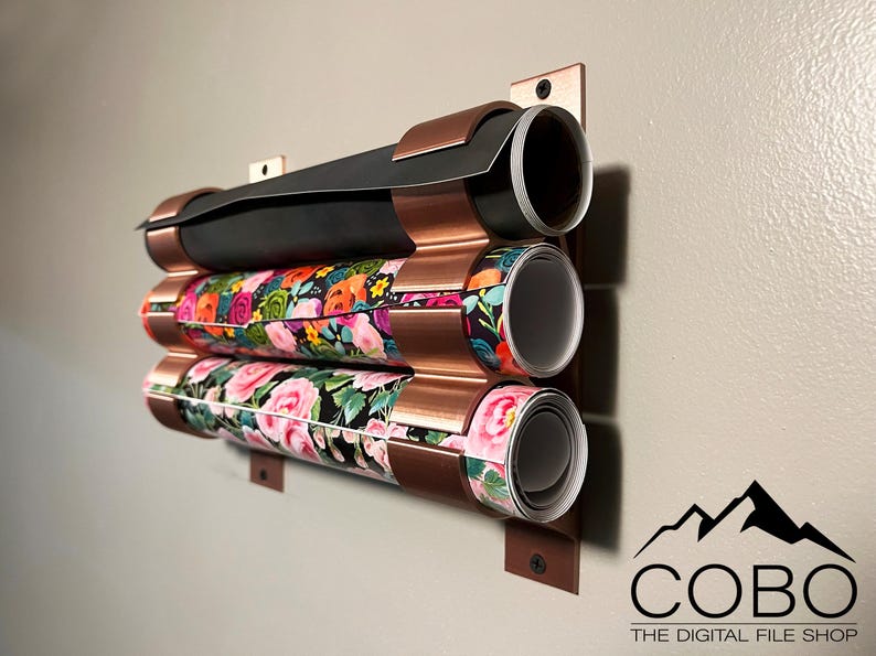 May include: A copper wall-mounted organizer with three rolls of patterned wrapping paper. The organizer has a corrugated design and is mounted to a wall with screws. The rolls of wrapping paper are in various floral patterns.
