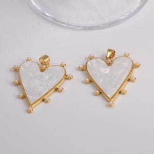 May include: Two gold-toned heart pendants with a white, pearlescent center. Each pendant is outlined in gold and adorned with small, round pearls. The pendants have a loop at the top for a chain.