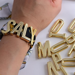 May include: A gold-colored bracelet spelling out "SALY" is wrapped around a wrist. The bracelet's letters are large and blocky. Additional gold letters are scattered on a white surface, suggesting customization options. The bracelet is on a patterned fabric.