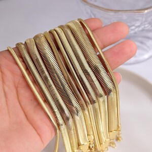 May include: A collection of gold-toned herringbone chains featuring a flat, flexible, and interlocking link design. The chains vary in width, displaying a smooth, reflective metallic surface with a consistent, repeating chevron pattern throughout.
