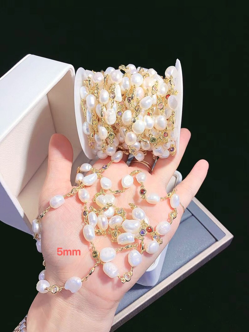 May include: A white pearl and gold chain with colorful gemstones. The chain is wound around a spool and is also laid out in a straight line. The pearls are 5mm in size.
