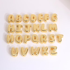 Può includere: Perline dorate dell'alfabeto, ogni lettera dell'alfabeto è rappresentata. Le perline sono disposte in file, con ogni lettera chiaramente visibile. Le perline hanno una finitura lucida e metallica.
