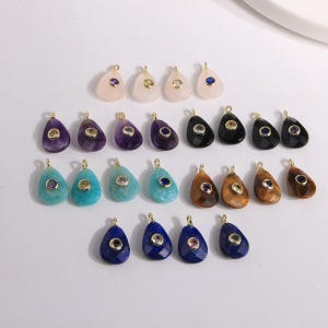 May include: A collection of teardrop-shaped gemstone pendants in various colors, including pink, purple, turquoise, brown, black, and blue. Each pendant has a gold-colored bail and a small, contrasting gemstone accent.