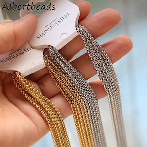 Può includere: Primo piano di più fili di catena dorata e argentata, etichettati "Stainless Steel". Le catene sono disposte a ventaglio e sono tenute in una mano.