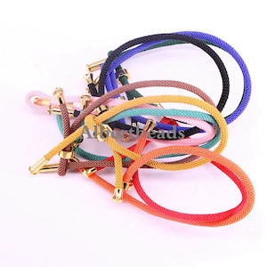 Könnte beinhalten: Eine Sammlung bunter Seil-Armbänder mit goldenen Metallverschlüssen. Die Armbänder sind in verschiedenen Farben, darunter rosa, orange, gelb, grün, blau und schwarz.