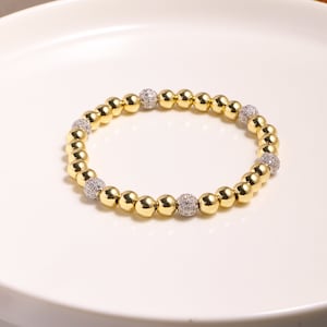 May include: A gold beaded bracelet with alternating smooth and textured beads. The textured beads have a sparkly finish.