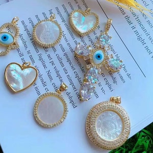 Può includere: Una collezione di nove ciondoli in oro con intarsi in madreperla bianca e iridescente. I ciondoli includono un cuore, un cerchio e un design di occhio malvagio. Alcuni ciondoli sono adornati con strass trasparenti.