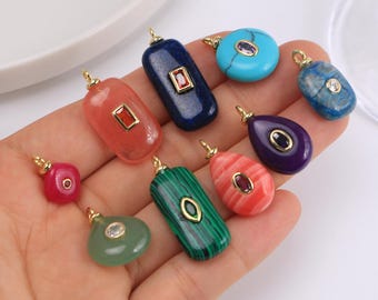 Puede incluir: Una colección de colgantes de piedras preciosas en varias formas y colores, incluyendo rojo, azul, verde y morado. Cada colgante tiene una fianza dorada y presenta una piedra preciosa o una incrustación decorativa. Los colgantes se sostienen en una mano.