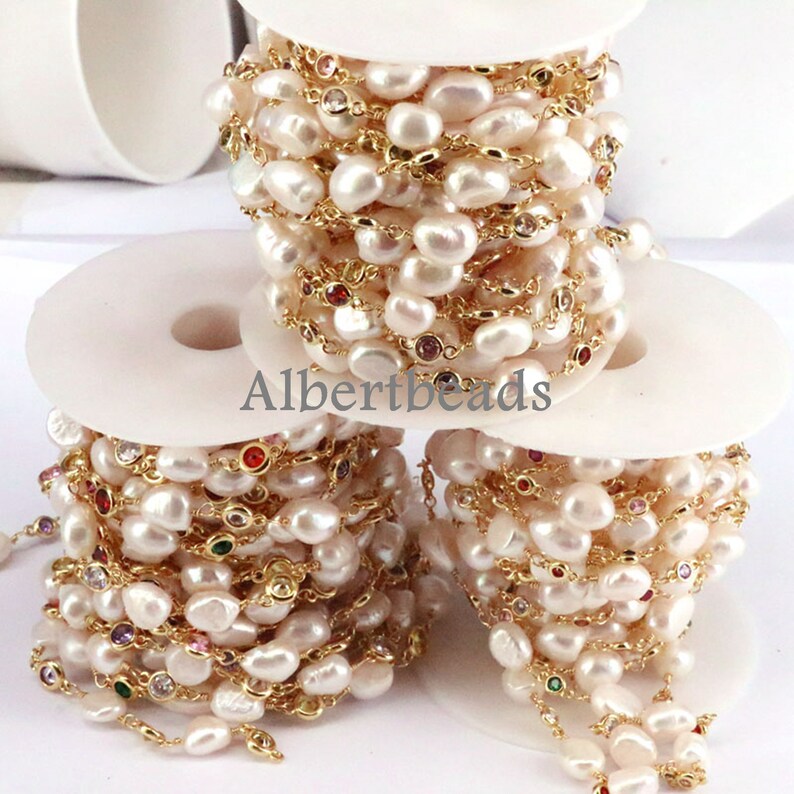 May include: A spool of gold chain with white baroque pearls and colorful faceted gemstones. The chain is wound around a white plastic spool. The text "Albertbeads" is printed on the spool.