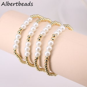 May include: Four gold and white beaded bracelets stacked on a wrist. The bracelets are made of small gold beads and larger white pearl-like beads.