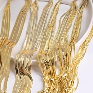 Puede incluir: Una colección de collares de cadena de espiga en tono dorado drapeados sobre una superficie circular blanca. Cada collar presenta un diseño de eslabones planos, flexibles y reflectantes que captan la luz, creando un brillo metálico.
