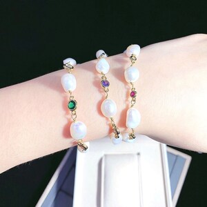 May include: Three gold pearl bracelets with colorful gemstone accents. The bracelets feature white pearls and small green, purple, and red gemstones.