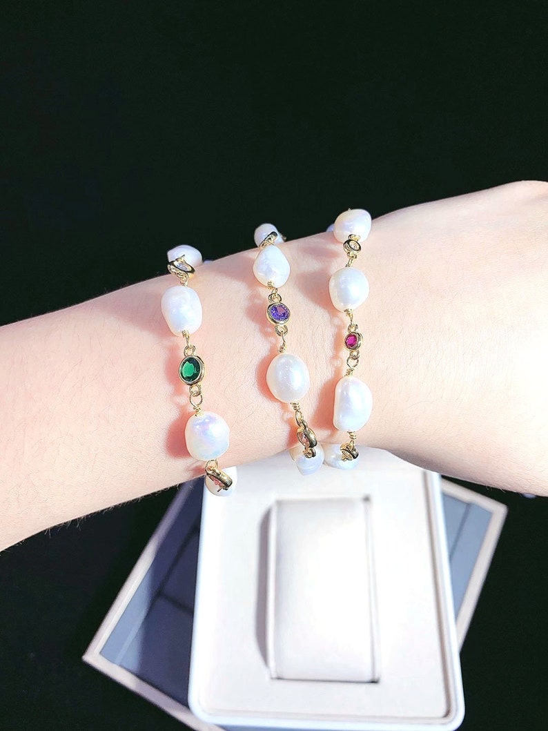 May include: Three gold pearl bracelets with colorful gemstone accents. The bracelets feature white pearls and small green, purple, and red gemstones.