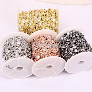 May include: A selection of decorative chain on spools, in gold, silver, rose gold, and black. Each chain features clear, round gemstones set in a delicate link design. The chains are wound on white spools, ideal for jewellery making.