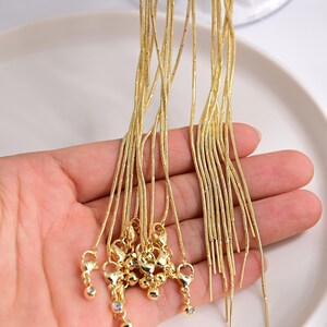 May include: A collection of multiple gold-toned snake chains, each featuring a lobster claw clasp at one end and a small decorative bead at the other. The chains are thin, flexible, and have a smooth, metallic texture throughout their length.