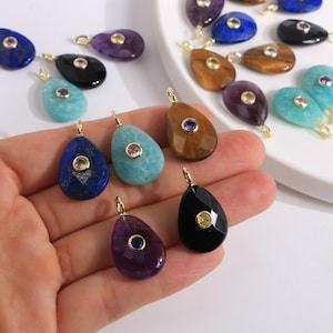 May include: A collection of teardrop-shaped gemstone pendants in various colors, including blue, purple, black, and brown. Each pendant features a smaller, contrasting gemstone in the center and a gold-colored bail for hanging. The gemstones are displayed on a white surface.