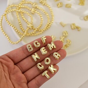 Peut inclure: Charm en forme d'alphabet de couleur or avec des accents de cristaux transparents. Les breloques sont en forme de lettres variées, dont B, G, C, F, E, M, N, O, A, T et K. Plusieurs colliers de perles dorées sont également visibles.