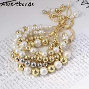 May include: A set of four bracelets with gold and white beads. The bracelets are made with a combination of round gold beads, white pearls, and silver beads. The bracelets are adjustable with a sliding knot.