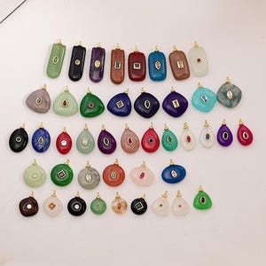 Puede incluir: Una colección de colgantes de piedras preciosas en varias formas y colores, incluyendo verde, negro, morado y rojo. Cada colgante tiene una argolla dorada y presenta una piedra preciosa o cristal. Los colgantes están dispuestos sobre una superficie blanca.