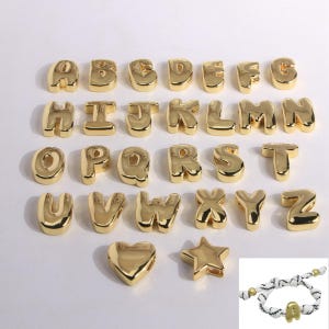 Puede incluir: Una colección de abalorios dorados con letras del alfabeto, cada uno con un diseño redondeado en forma de burbuja. Las letras están dispuestas en filas, con un charm de corazón y estrella debajo. También se muestra una pulsera con un charm de letra dorada.