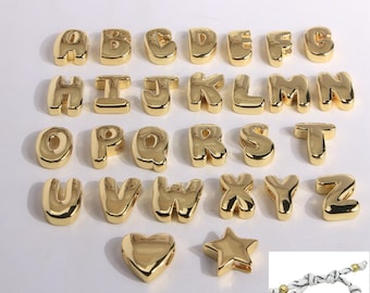 May include: A collection of gold-colored alphabet letter charms, each with a rounded, bubble-like design. The letters are arranged in rows, with a heart and star charm below. A bracelet with a gold letter charm is also shown.