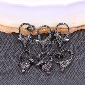 May include: Six black metal lobster clasps with clear cubic zirconia stones. The clasps are oval shaped and have a spring-loaded closure.