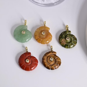 Op de afbeelding: Vijf edelsteenhangers in de vorm van een spiraalvormige schelp. Elke hanger heeft een goudkleurige beugel en een kleine, ronde, heldere steen in het midden. De edelstenen zijn groen, oranje, groen met bruin, rood en bruin.