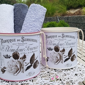 Könnte beinhalten: Zwei weiße Metallbehälter mit rosa und cremefarbenen Deckeln und Seilgriffen. Die Behälter haben ein Vintage-Blumendesign mit dem aufgedruckten Text "Fabrique de Serrurerie pour Balingerts Slomdeur: Tournier 12 Rue de la Madeleine". Die Behälter sind mit Handtüchern gefüllt.
