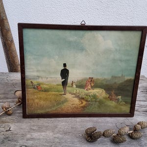 Könnte beinhalten: Gerahmtes Vintage-Landschaftsgemälde mit dunklem Holzrahmen. Das Kunstwerk zeigt einen Mann in schwarzem Mantel und Zylinder, der auf einem Pfad steht und eine malerische Aussicht mit Figuren und Gebäuden überblickt. Das Gemälde hat eine gedämpfte Farbpalette.