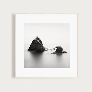Puede incluir: Una fotografía en blanco y negro de un puente de piedra que conecta dos islas rocosas. El puente está hecho de piedra y madera y está rodeado de agua. El agua está tranquila y quieta, y el cielo está nublado.