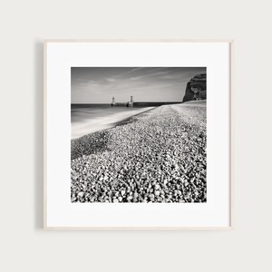 May include: Black and white photograph of a rocky beach with a pier and two small buildings in the distance. The water is blurred in the foreground, creating a sense of motion.