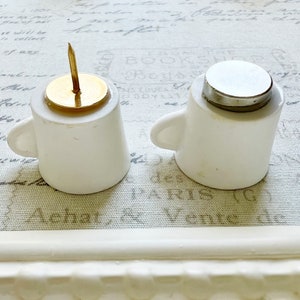 May include: Two white ceramic cups with lids. One cup has a gold top with a small, gold pin. The other cup has a silver lid.