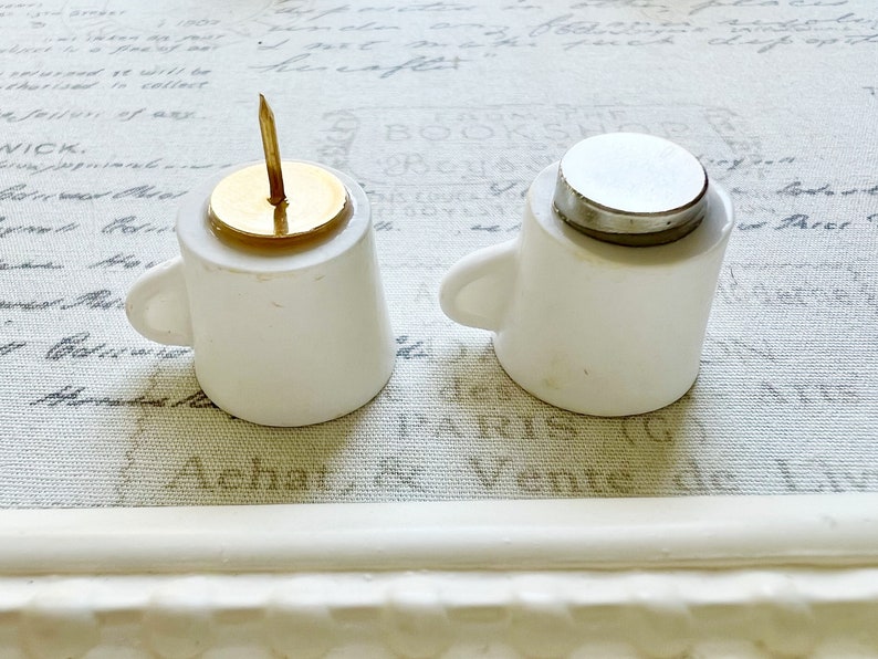 May include: Two white ceramic cups with lids. One cup has a gold top with a small, gold pin. The other cup has a silver lid.