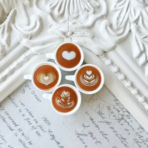 May include: Four miniature white ceramic cups filled with brown coffee and white foam designs. The cups are arranged in a circle on a white surface with handwritten text.