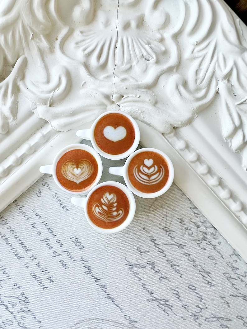 May include: Four miniature white ceramic cups filled with brown coffee and white foam designs. The cups are arranged in a circle on a white surface with handwritten text.