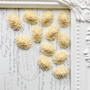 May include: Ten off-white oval-shaped resin cabochons with a floral design. The cabochons are arranged in a semi-circle on a white surface with a floral pattern.